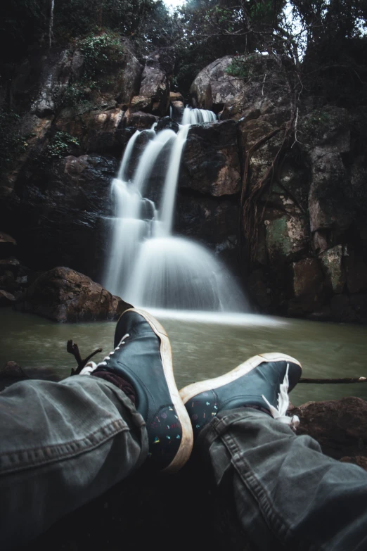 person's feet resting on the ground in front of a waterfall