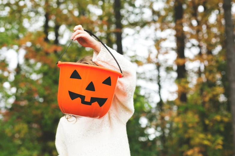 the woman is dressed in a white ponc, and holding a plastic bucket with a pumpkin face