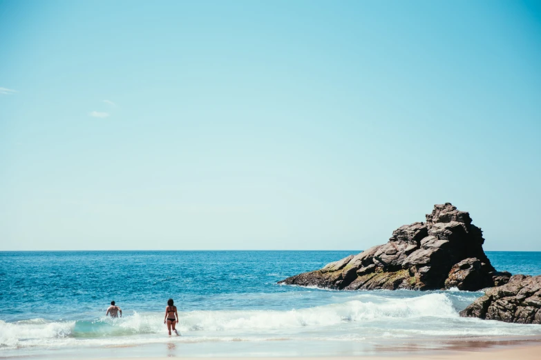 two people wading through the ocean with a rock formation in the background