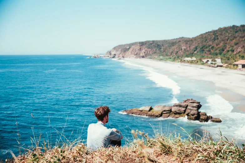 a person sitting at the edge of a cliff looking over a beautiful beach