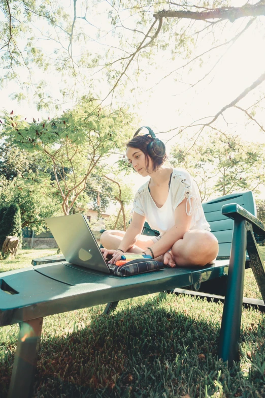 a woman wearing headphones sitting on a park bench working on her laptop
