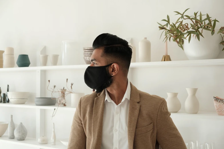 a man in a suit stands wearing a black face mask in front of the wall filled with ceramics