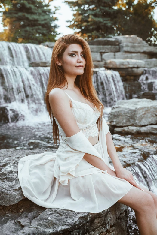 a woman with long red hair sitting on rocks by a waterfall