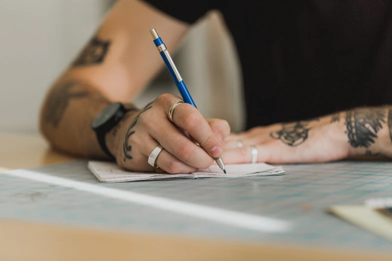 tattooed man with a ring sits at a desk and writes on paper