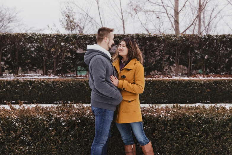 a woman hugging the arm of her boyfriend standing next to a hedges