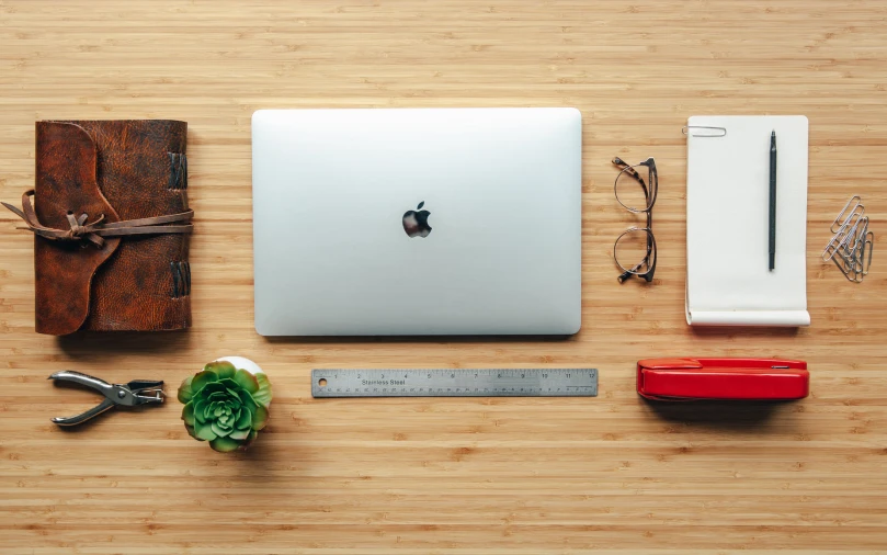 a computer, pen, ruler, scissors and leather journal on a wooden table