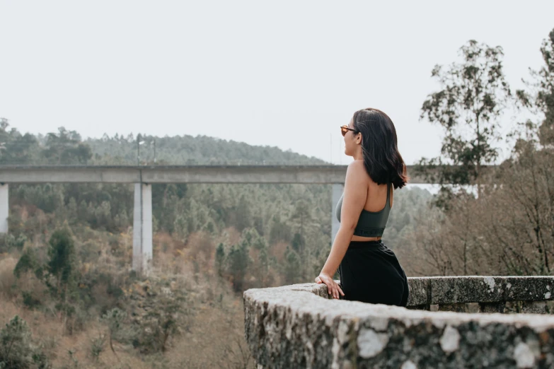 a woman with long dark hair sitting on a stone wall overlooking a bridge