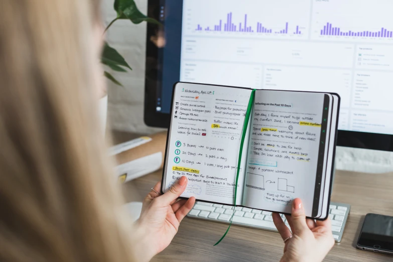 a woman with a notebook open sitting in front of a monitor