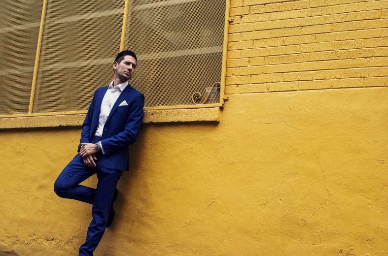 a man in a blue suit leaning against a yellow wall