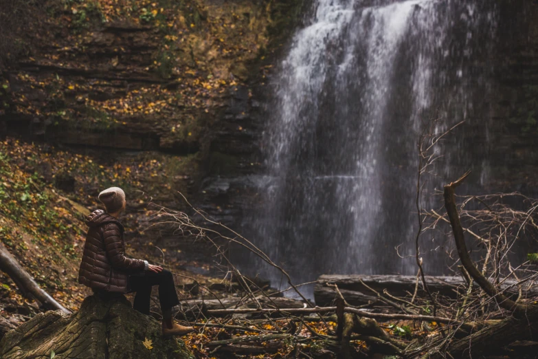 a person sitting on top of a log near a tall waterfall