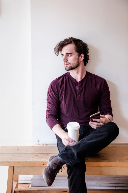 man in maroon shirt sitting on bench holding a coffee