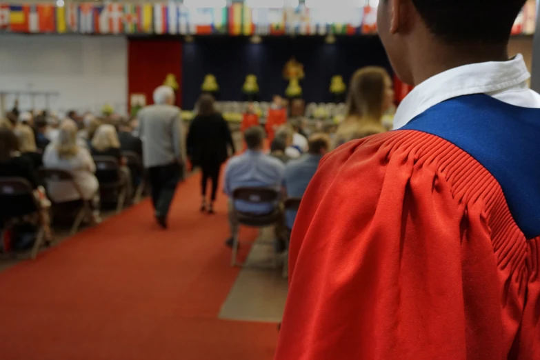 man in red and blue graduation gown watching people in background