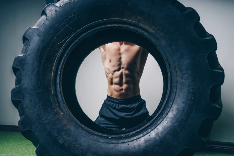 a man with  on, without a shirt, standing behind an oversized tire