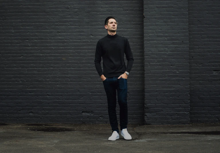 a man in black shirt and jeans stands in front of a brick wall