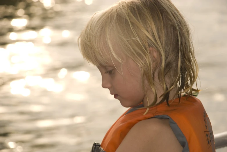 a little girl with blonde hair and an orange life vest looking out over water