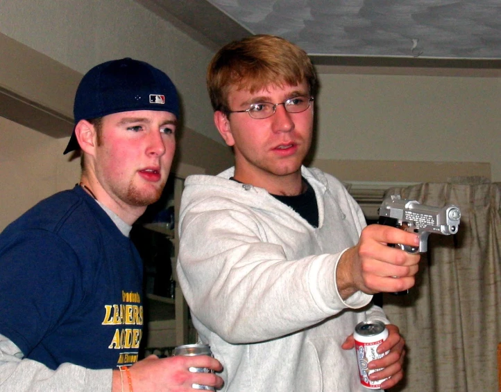 two boys standing together in a room holding drinks and controllers