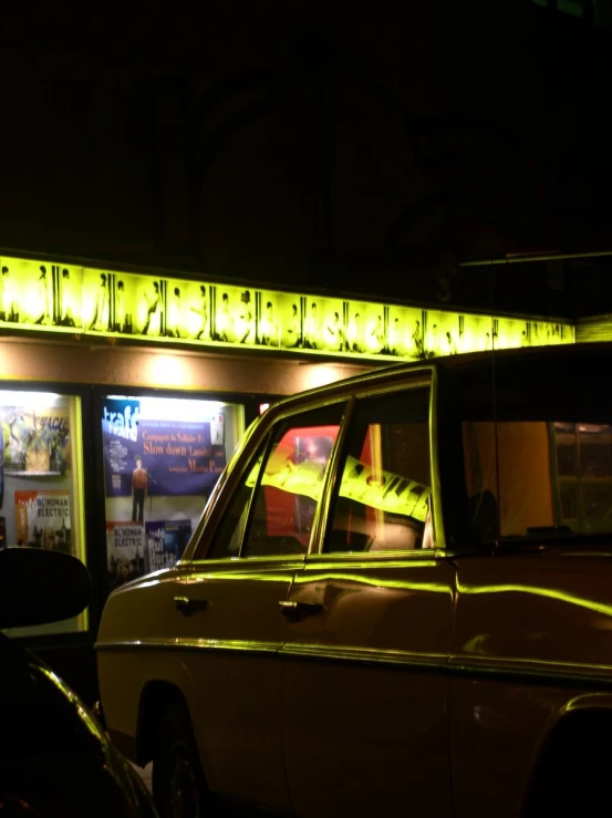 a yellow car sits outside a neon sign shop