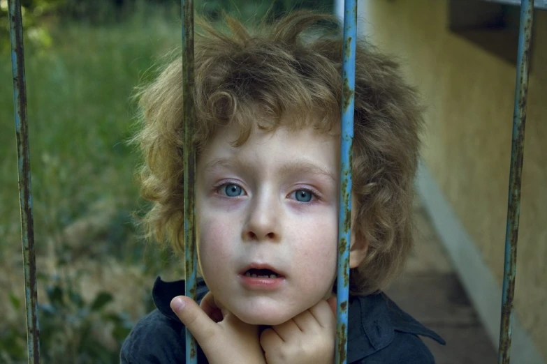 a boy with curly hair leans on a fence