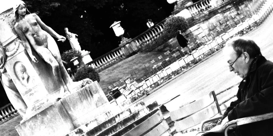 black and white pograph of man sitting on park bench near stone statues