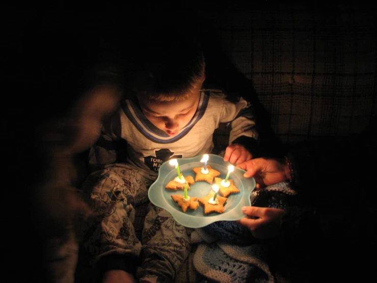 a boy sitting in the dark holding a plate of cookies with lit candles