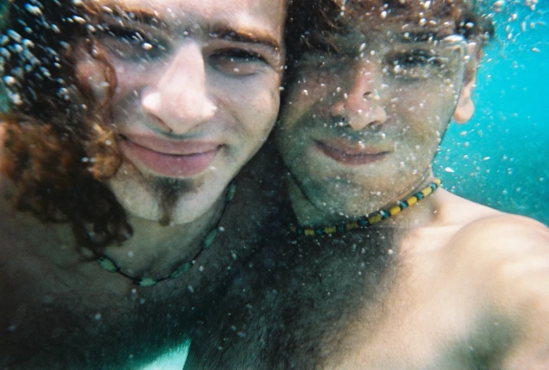 two men, one man with beard and necklace, swim underwater in a pool