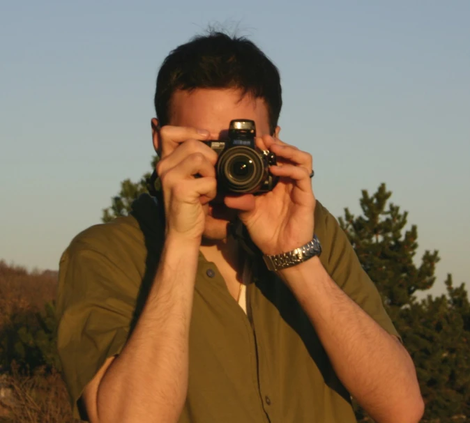 man holding camera in front of his face with trees in background