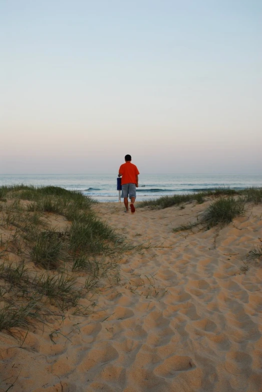 a man in red shirt walking up a sand dune near the ocean