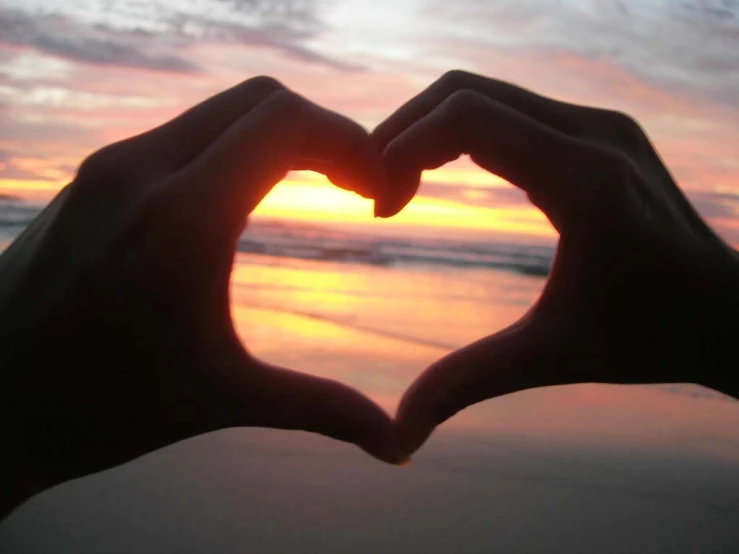 a hand making a heart shape with the sun setting in the background