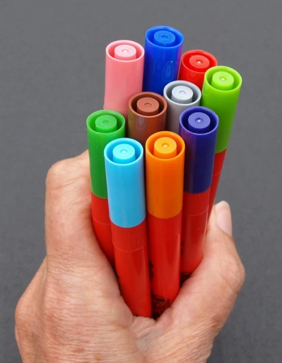 small hand holding colorfully painted markers next to gray background