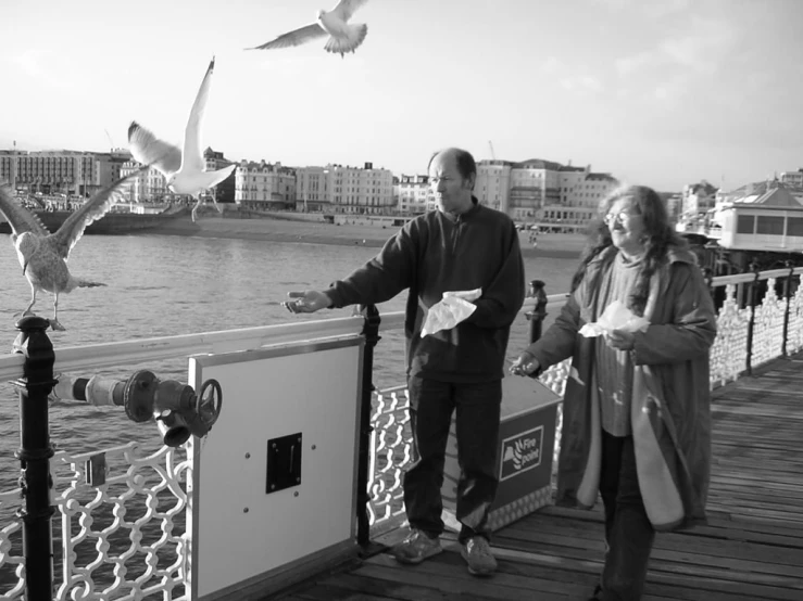 man and woman standing on a bridge near water while seagulls fly overhead