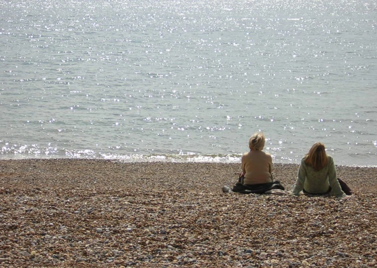 two people sitting on the beach while looking at the water