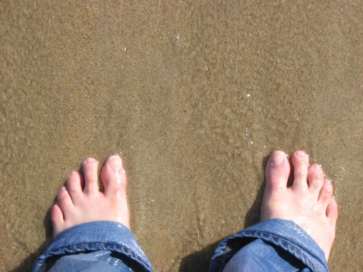 someones feet are standing on the beach and a surfboard in the background