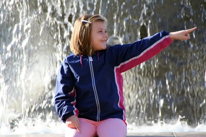 a little girl sitting on the ground pointing at a fountain