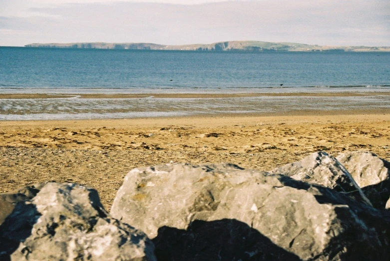 large rocks are on the beach, and a lone surfer is walking across the water in the distance