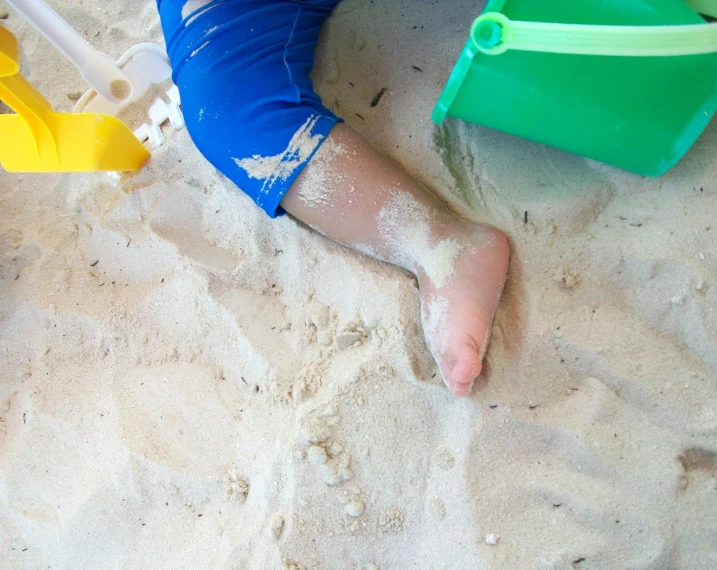 sand with the feet of a person in blue shorts and yellow plastic shovels