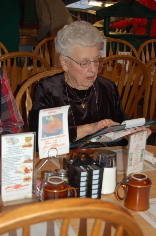 an elderly woman reading a magazine at a table full of tea and coffee