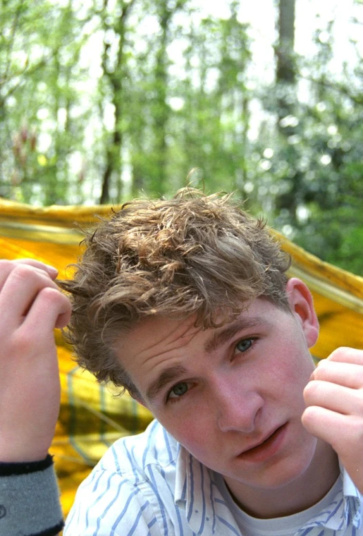 a boy holds up his hair while standing near the woods