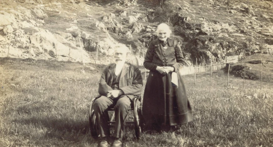 two old people in historical dress sitting on chairs outside