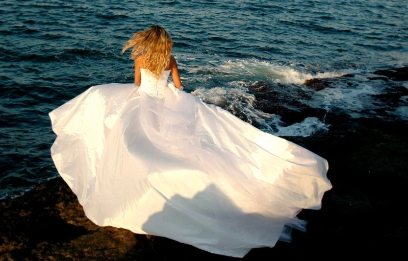 a woman wearing a white gown walking along a shore