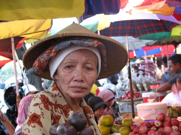 a woman with an umbrella hat standing in front of a fruit stand