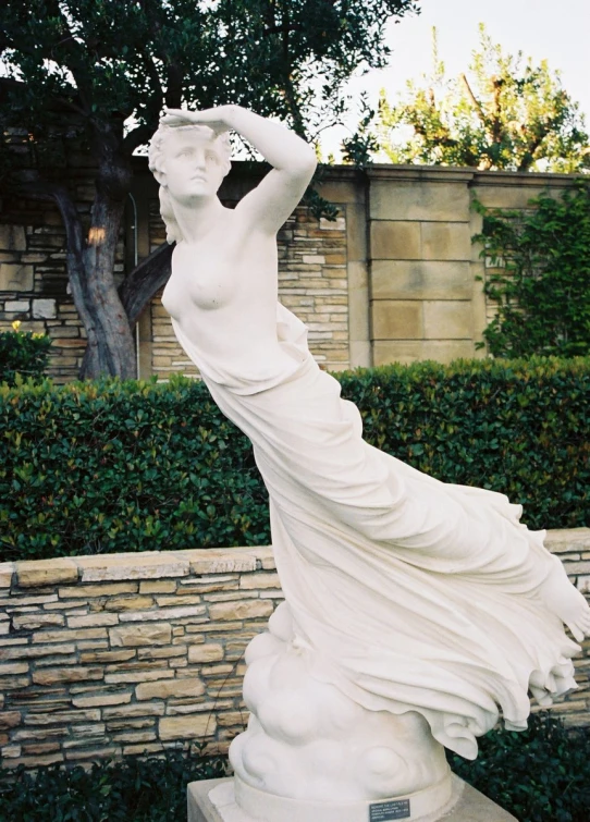 a statue of a woman wearing a long flowing white dress is in the foreground and bushes on either side