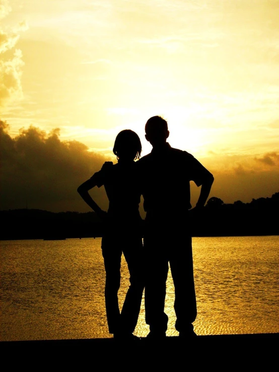 two people standing on the edge of a body of water while the sun sets