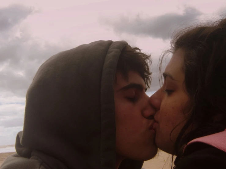 two people wearing hoods kiss while standing near the ocean