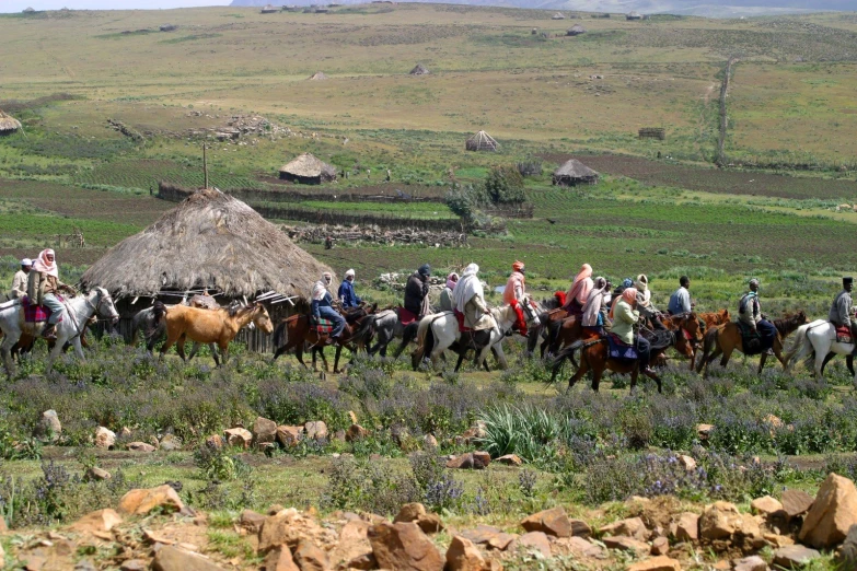 several people ride horses with some thatched hut in the background