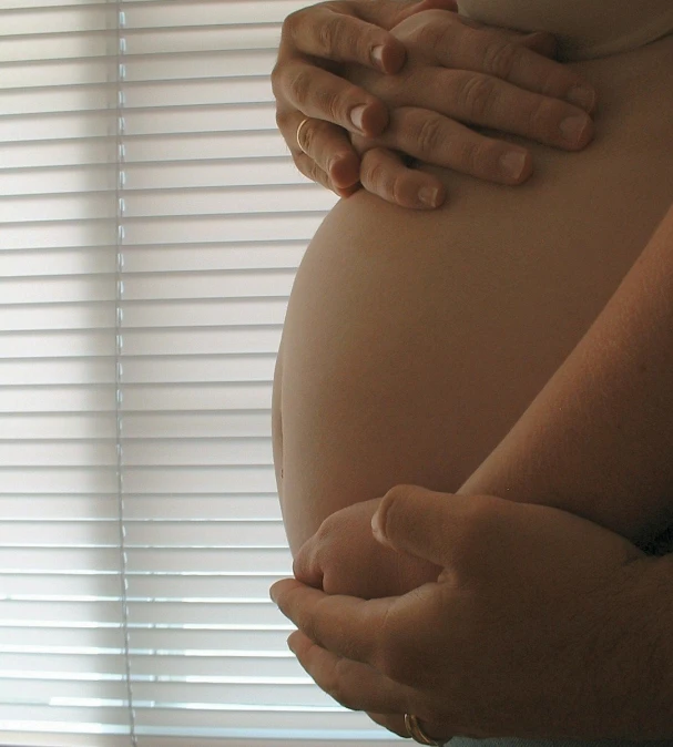 a pregnant person in front of blinds holds their hand on the belly