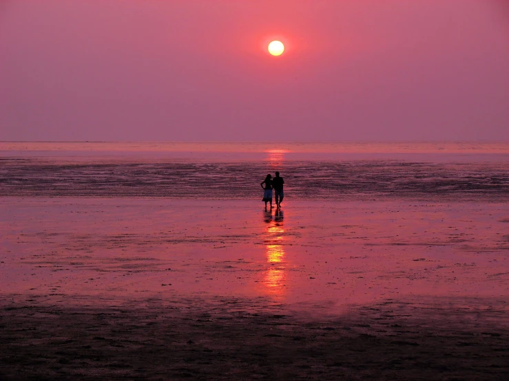 two people walking on the beach at sunset with the sun setting in the distance