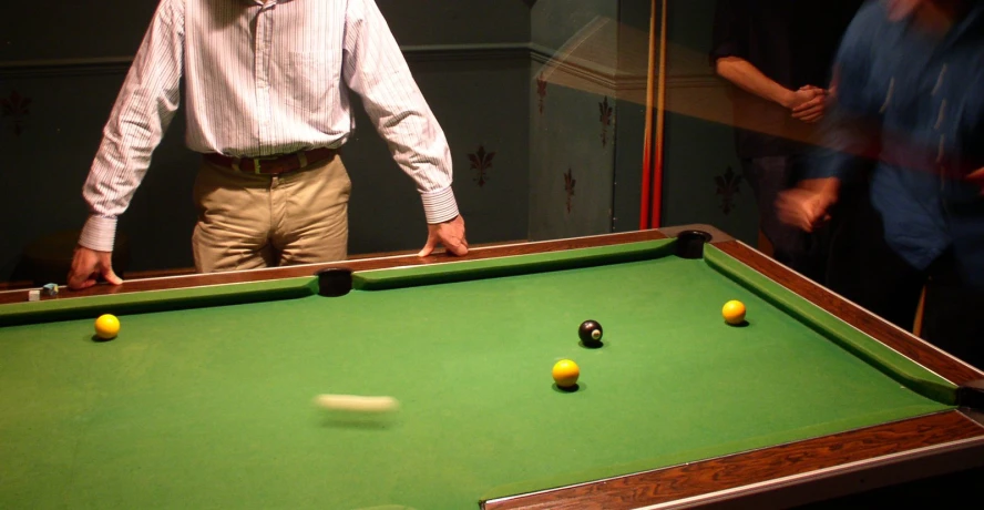 an adult standing at a pool table and a man in a blue shirt is leaning on the pool table