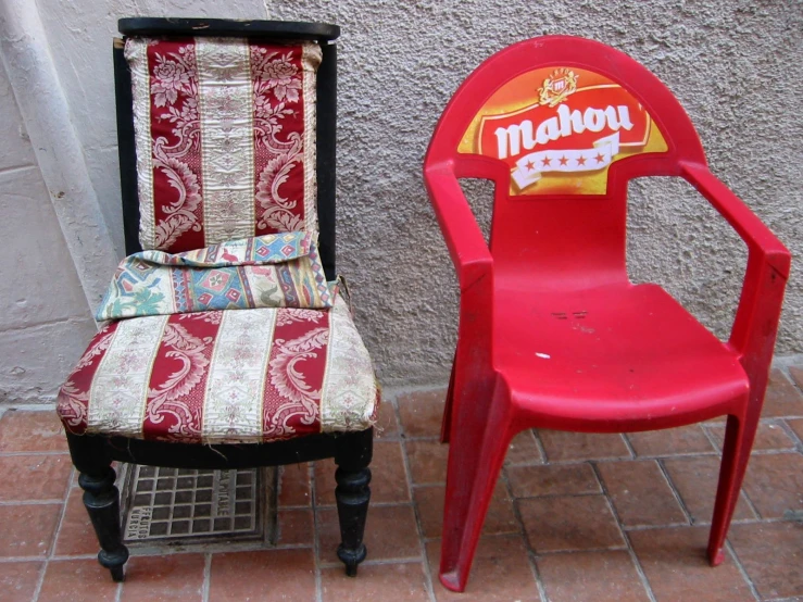 an empty red plastic chair and a miniature piece of furniture with patches of fabric on them, both sitting side by side