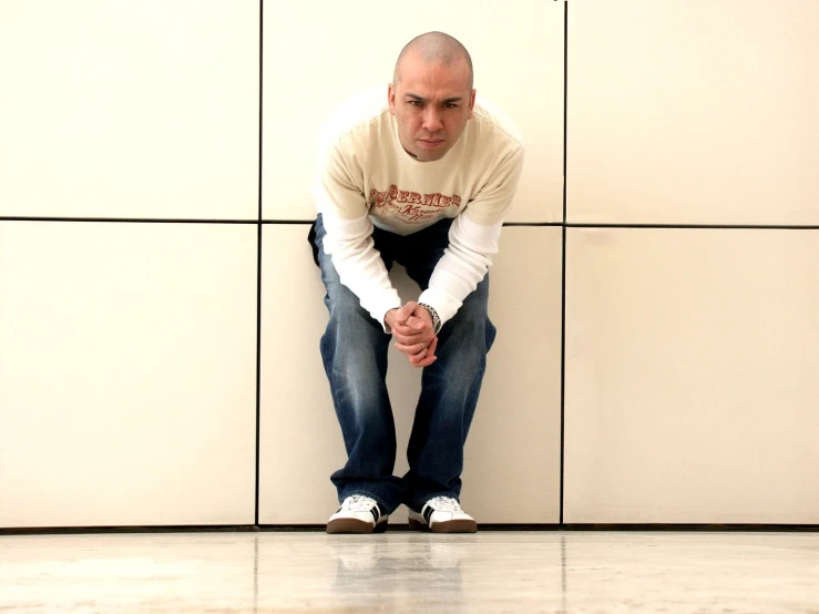 a man wearing jeans and a shirt sitting against a wall