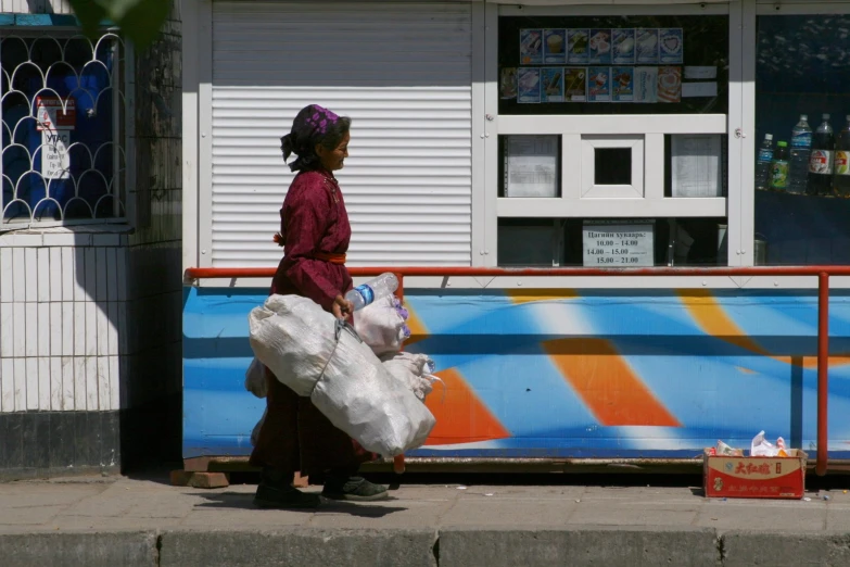 a woman carrying bags walks down a sidewalk in front of an outdoor convenience store